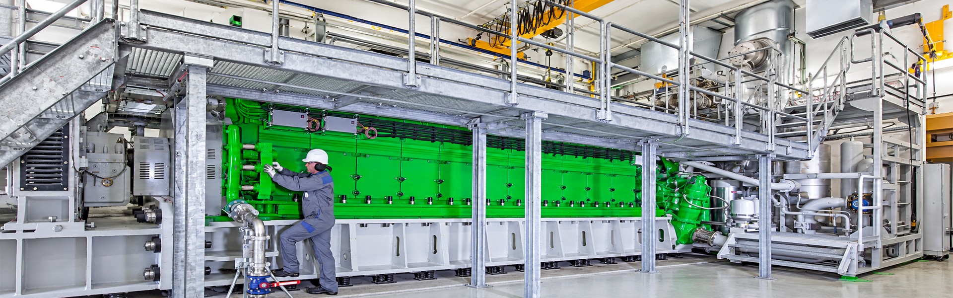 Technician inspecting a large green industrial engine beneath a galvanized maintenance platform.