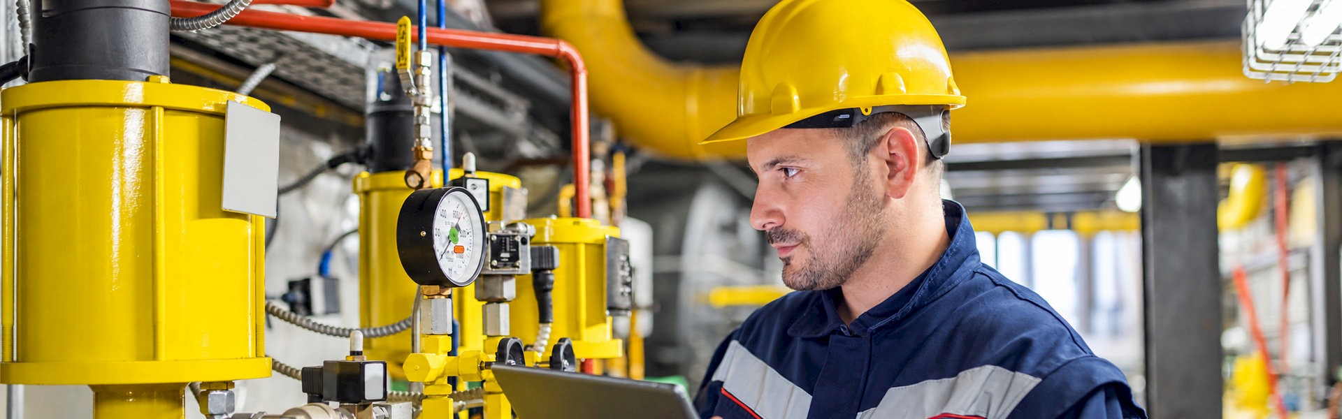 Technician holding a tablet while inspecting a pressure gauge and industrial process equipment.