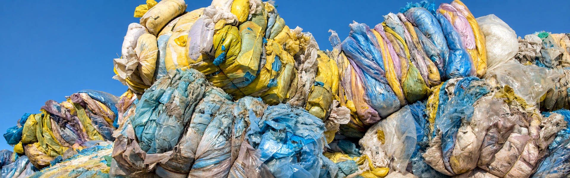 Stacked bales of compressed yellow, blue and clear plastic film awaiting recycling beneath a blue sky.