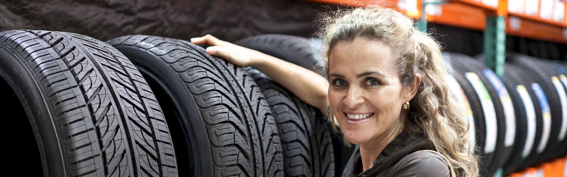Smiling woman resting her hand on a tire beside rows of tires in a warehouse.