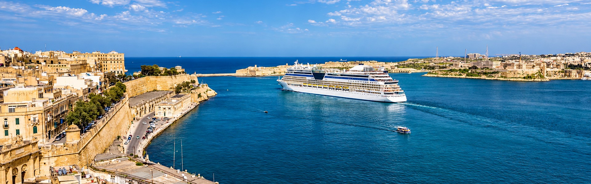 Cruise ship sailing through Malta’s Grand Harbour beside historic limestone buildings and waterfront fortifications.