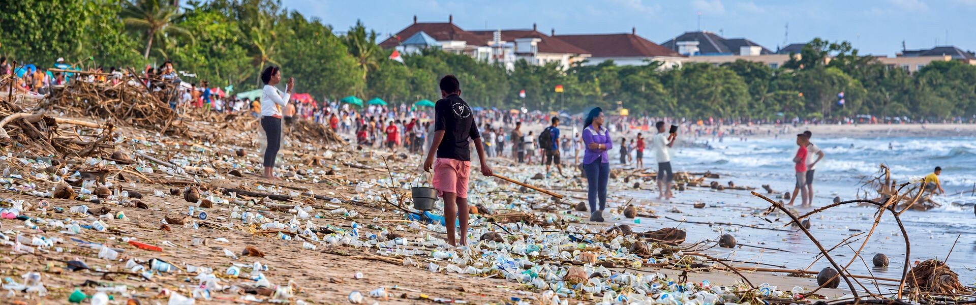 People along a beach covered with discarded plastic bottles, packaging and driftwood beside the ocean.
