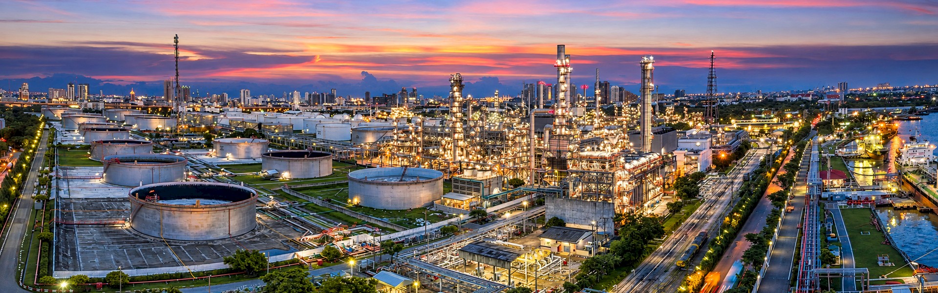 Industrial processing complex at sunset with illuminated towers, storage tanks and pipework beside a waterway.