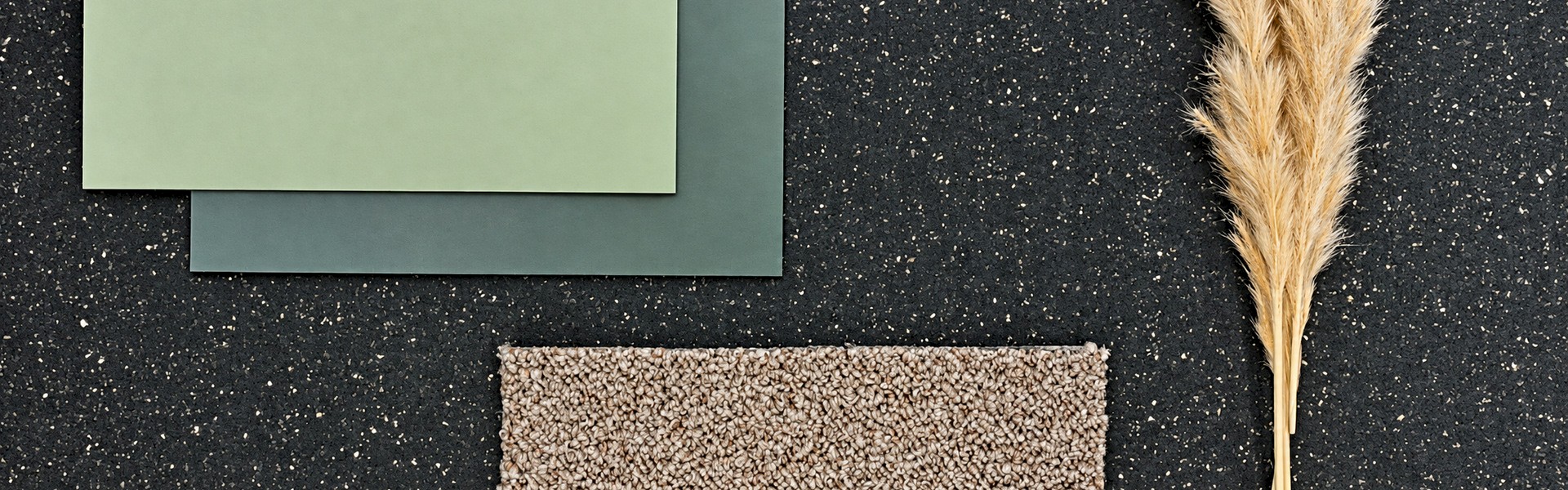 Green flooring samples and a beige carpet tile on a speckled black rubber surface beside dried pampas grass.