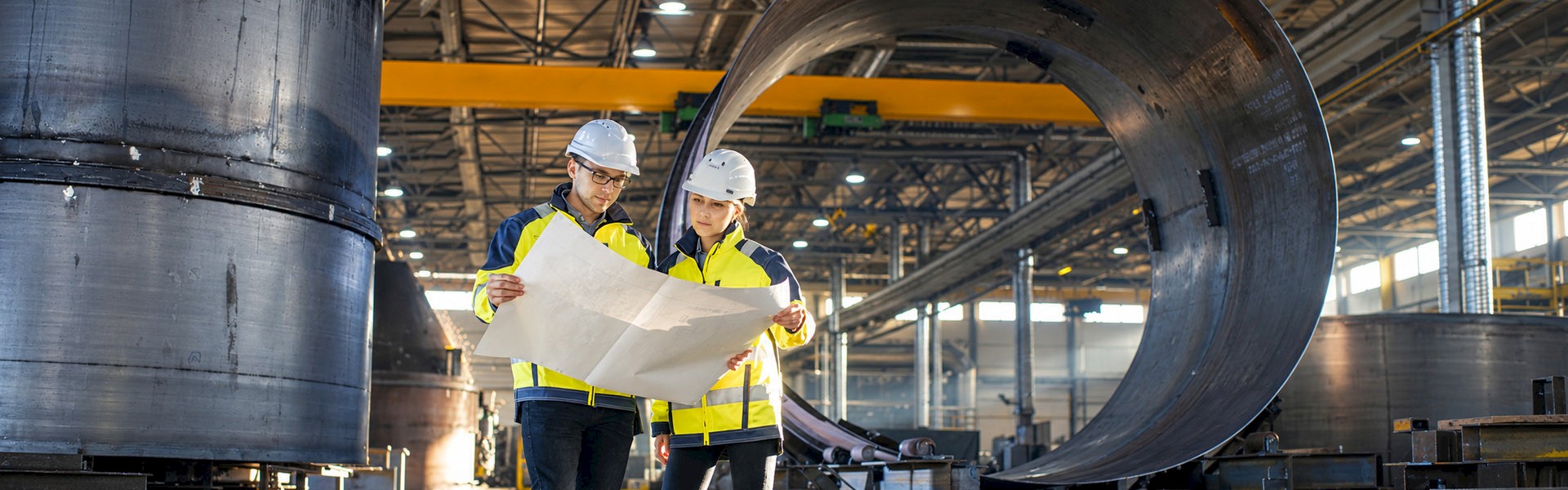 Two engineers reviewing technical drawings beside large cylindrical steel components in a fabrication workshop.